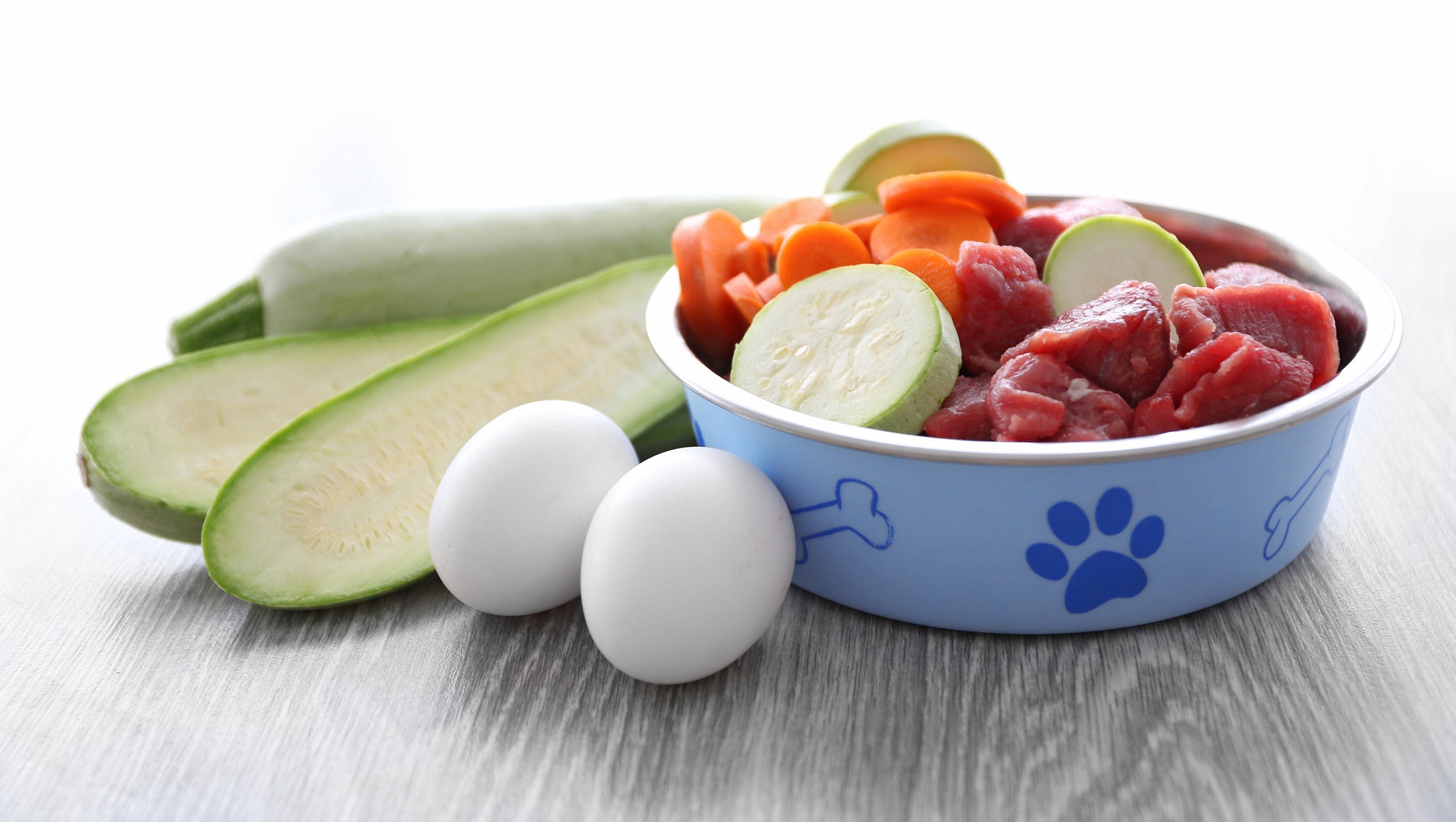 Blue dog bowl filled with meat and vegetables beside sliced zucchini and eggs