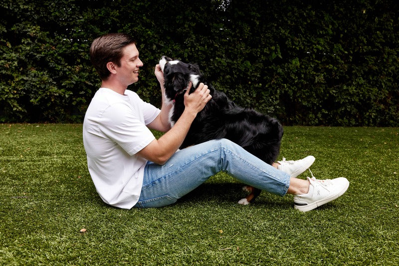Man sitting on grass and petting a black and white dog outdoors